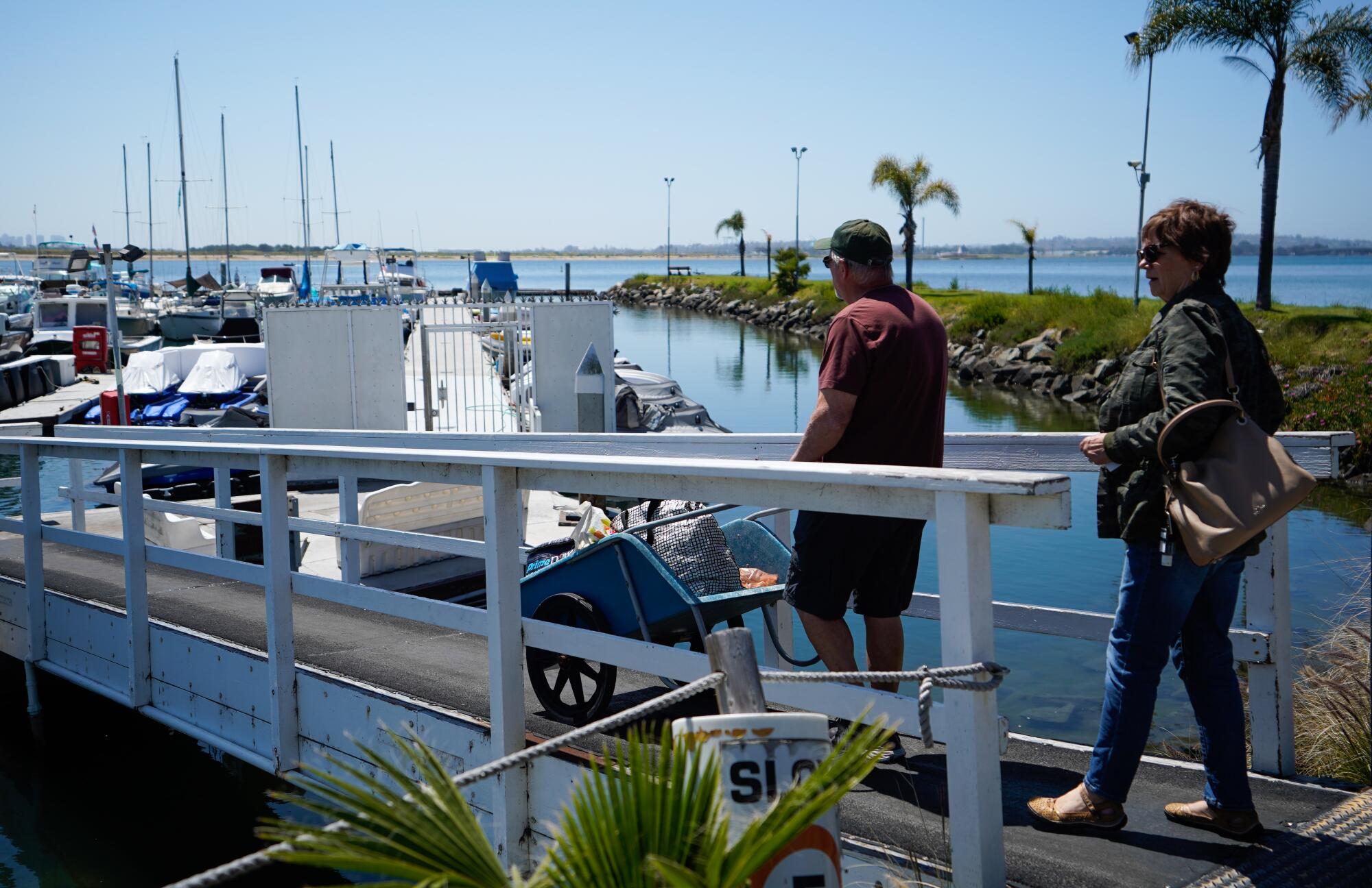 Two people walk across a wooden pedestrian bridge onto a dock at a marina flanked by palm trees.