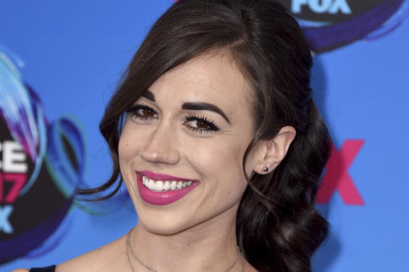 Colleen Ballinger with bangs and a ponytail wearing a scrappy black dress against a blue backdrop