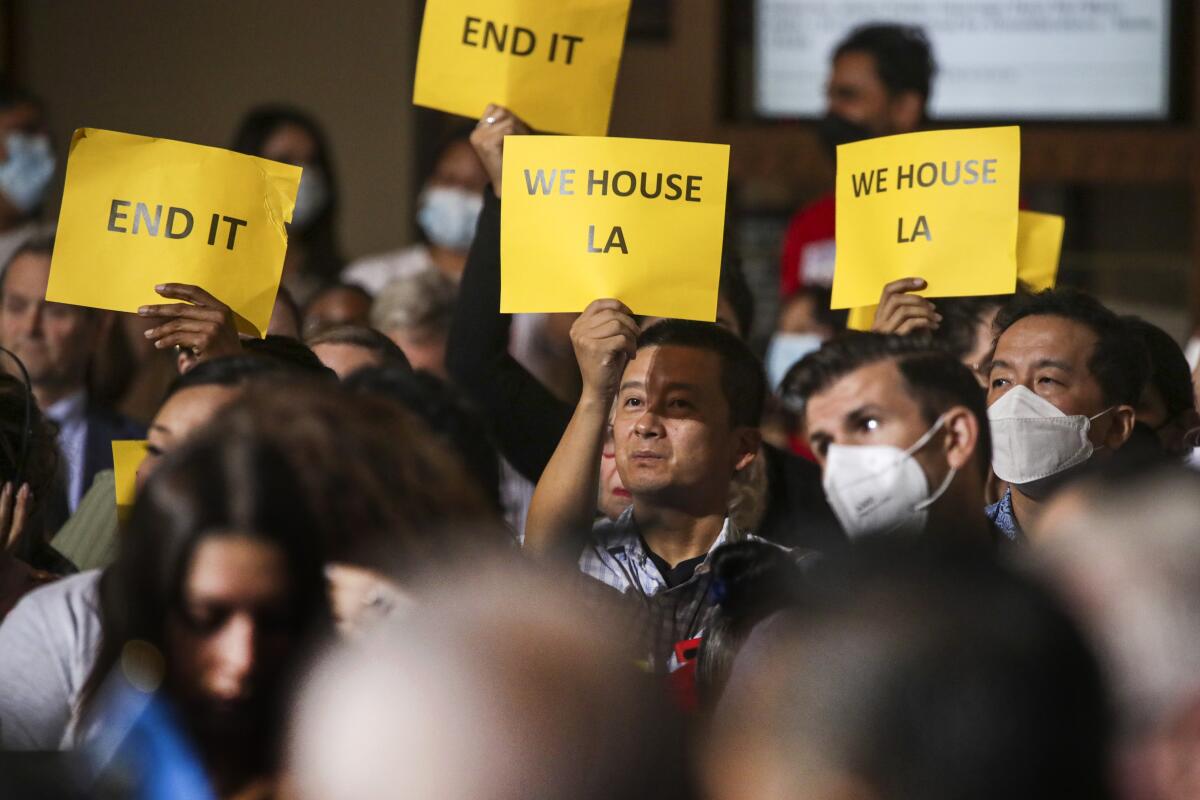 In a crowd of people, some in masks, several people lift small posters that say "We house L.A." and "End it."