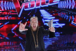 A man with long, blonde braids in a black suit holding out a rock sign and a metal trophy standing over confetti