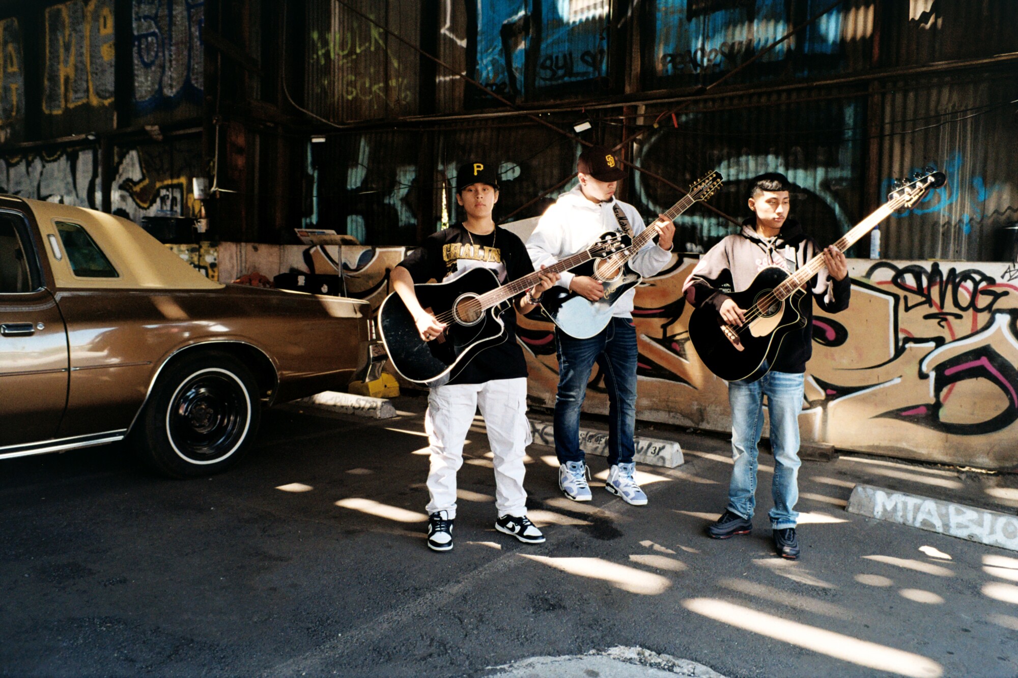 Three people stand with guitars near a vintage car in downtown Los Angeles.