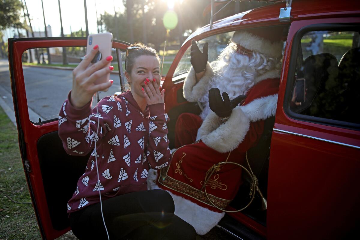A woman crouches and takes a selfie with a Santa sitting in the driver's seat of a red car