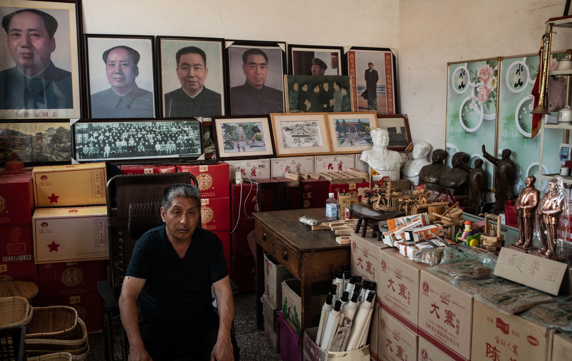 A man sits in shop where portraits of men and boxes with statues on top line the walls 