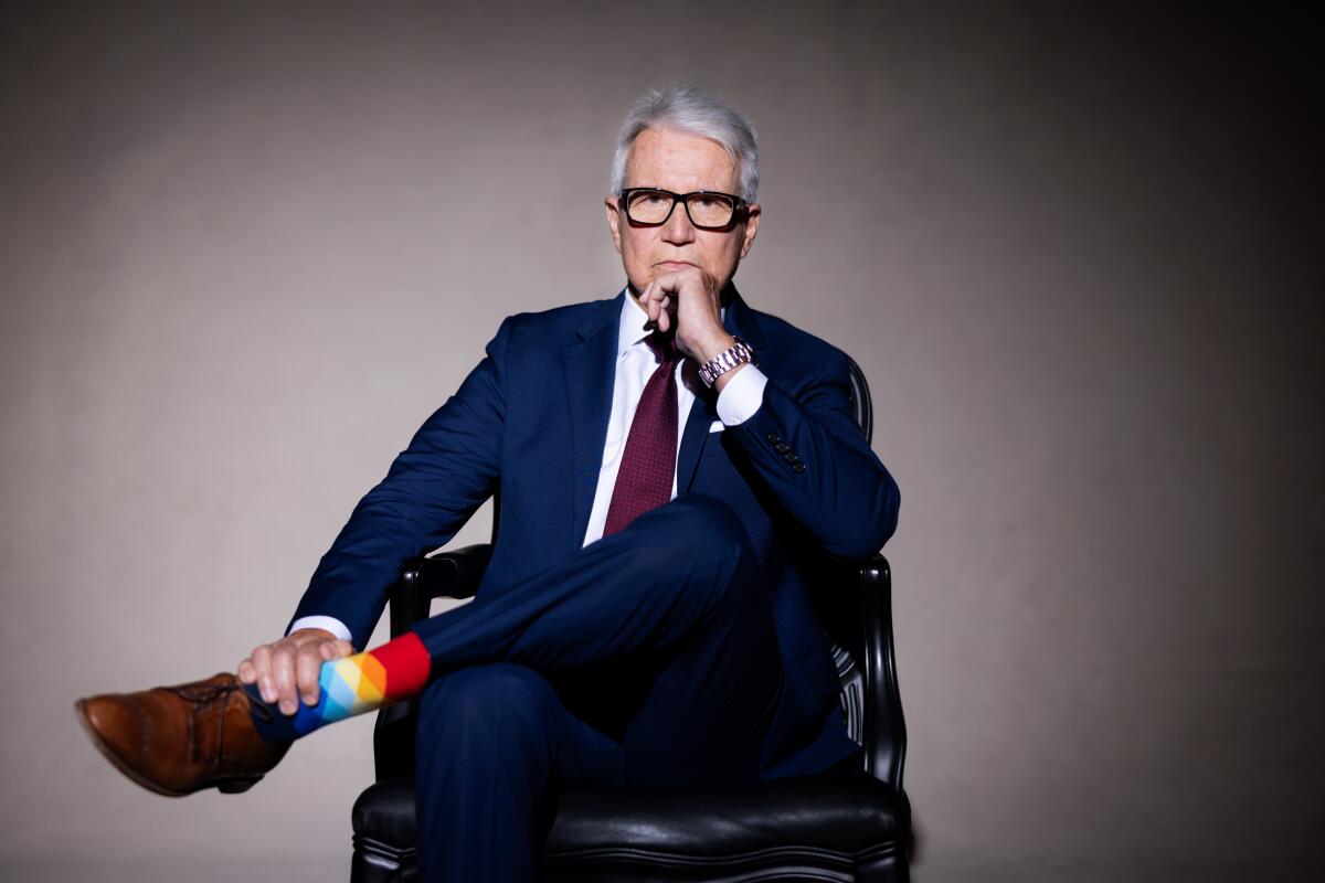 George Gascón, a gray-haired man in square glasses, navy suit and colorful argyle socks, poses unsmiling, legs crossed.
