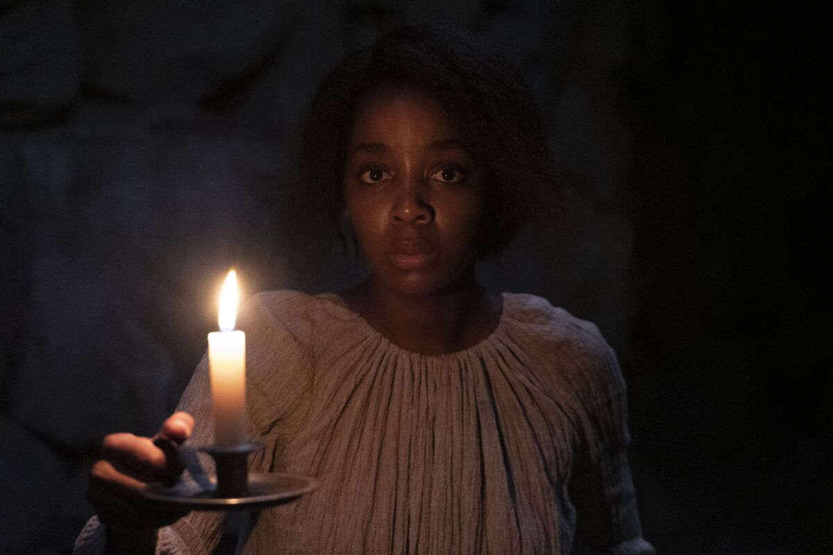 Thuso Mbedu, in a loose white gown, lifts an antique holder with a lighted candle in the dark.