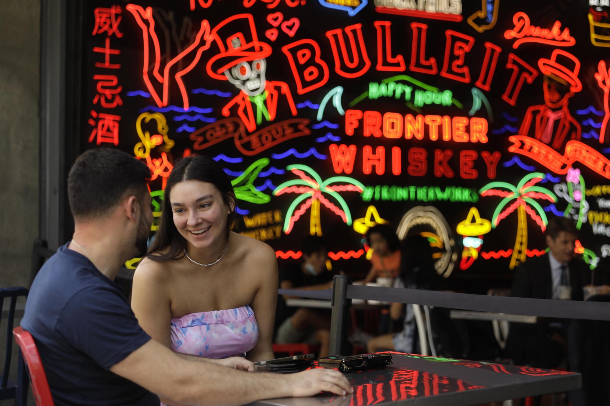 Two people talk in front of a large neon sign