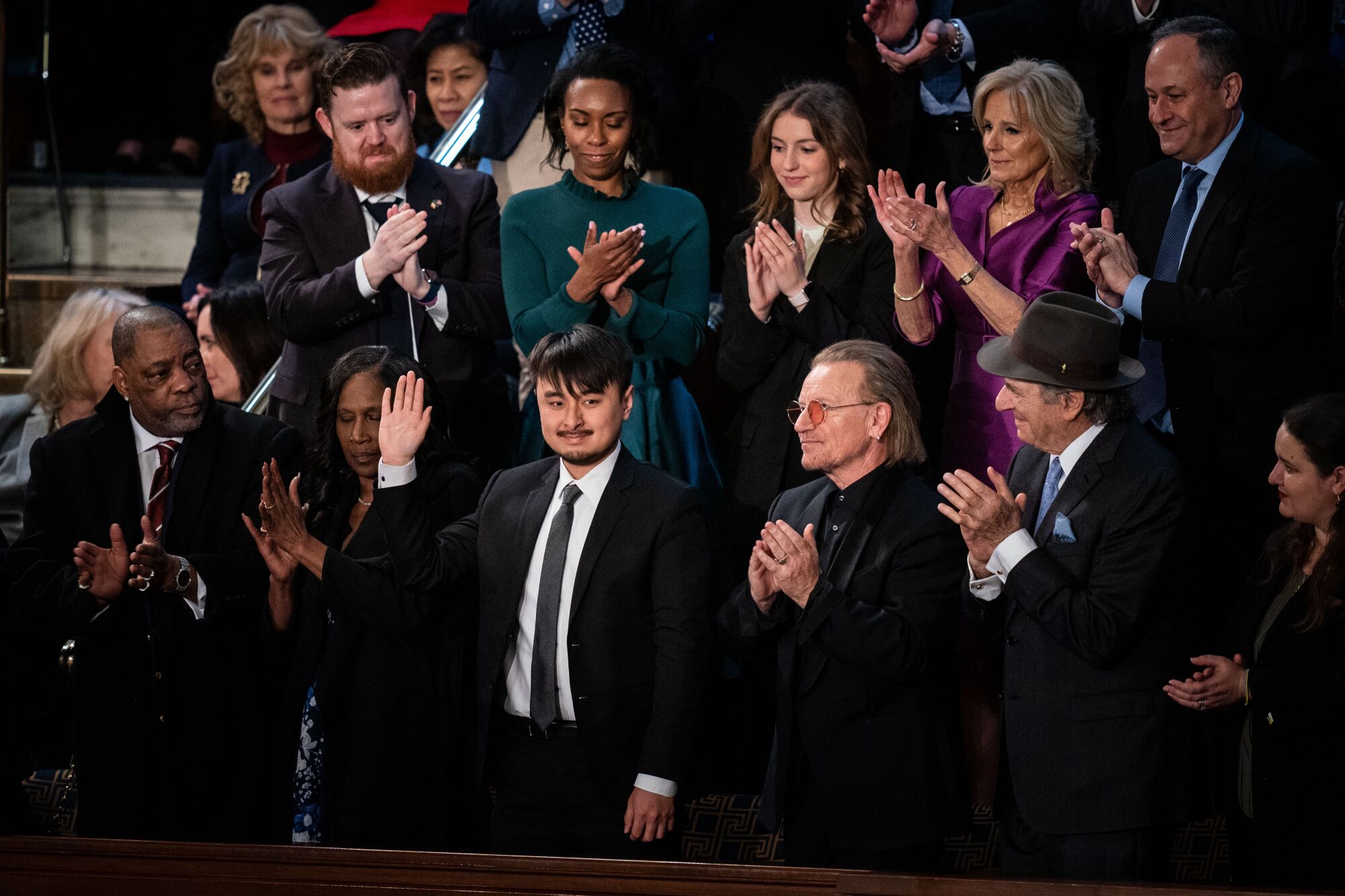 Un groupe de personnes bien habillées applaudit un jeune homme au milieu alors qu'il lève la main droite en guise de salutation.