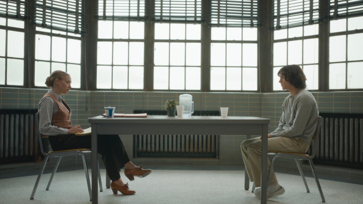 A woman sits across a long table from a man in a room filled with windows.