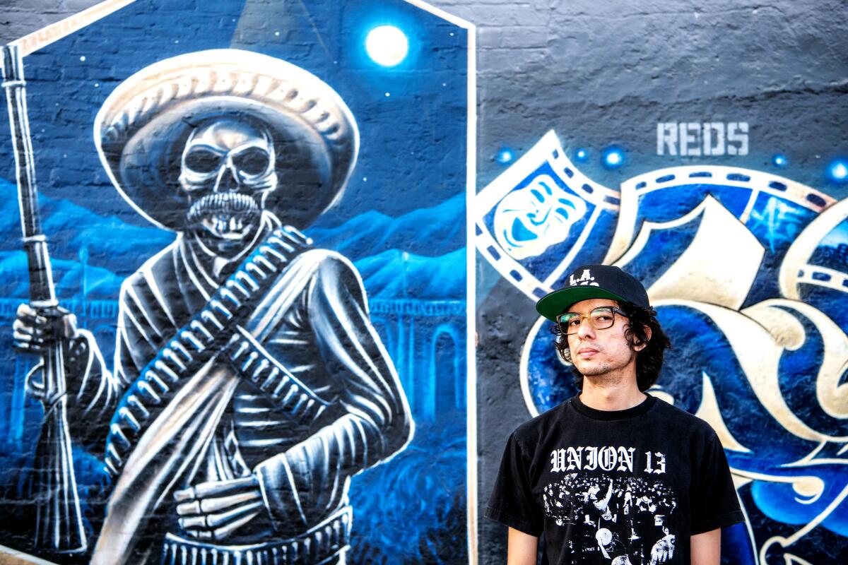A man in a black hat and a black shirt that says "Union 13" stands next to a mural of a skeleton in a sombrero with a rifle.