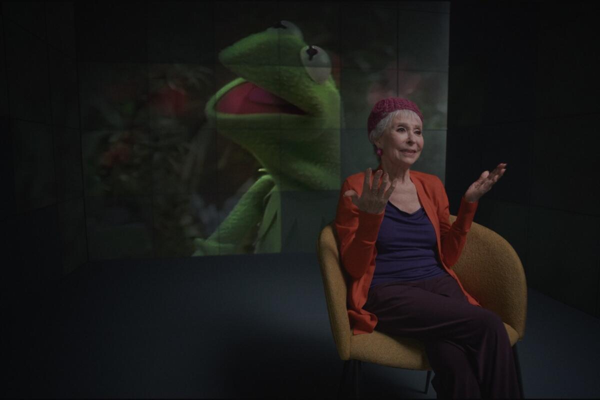 An image of Kermit the Frog on a giant screen behind Rita Moreno sitting in a chair gesturing.