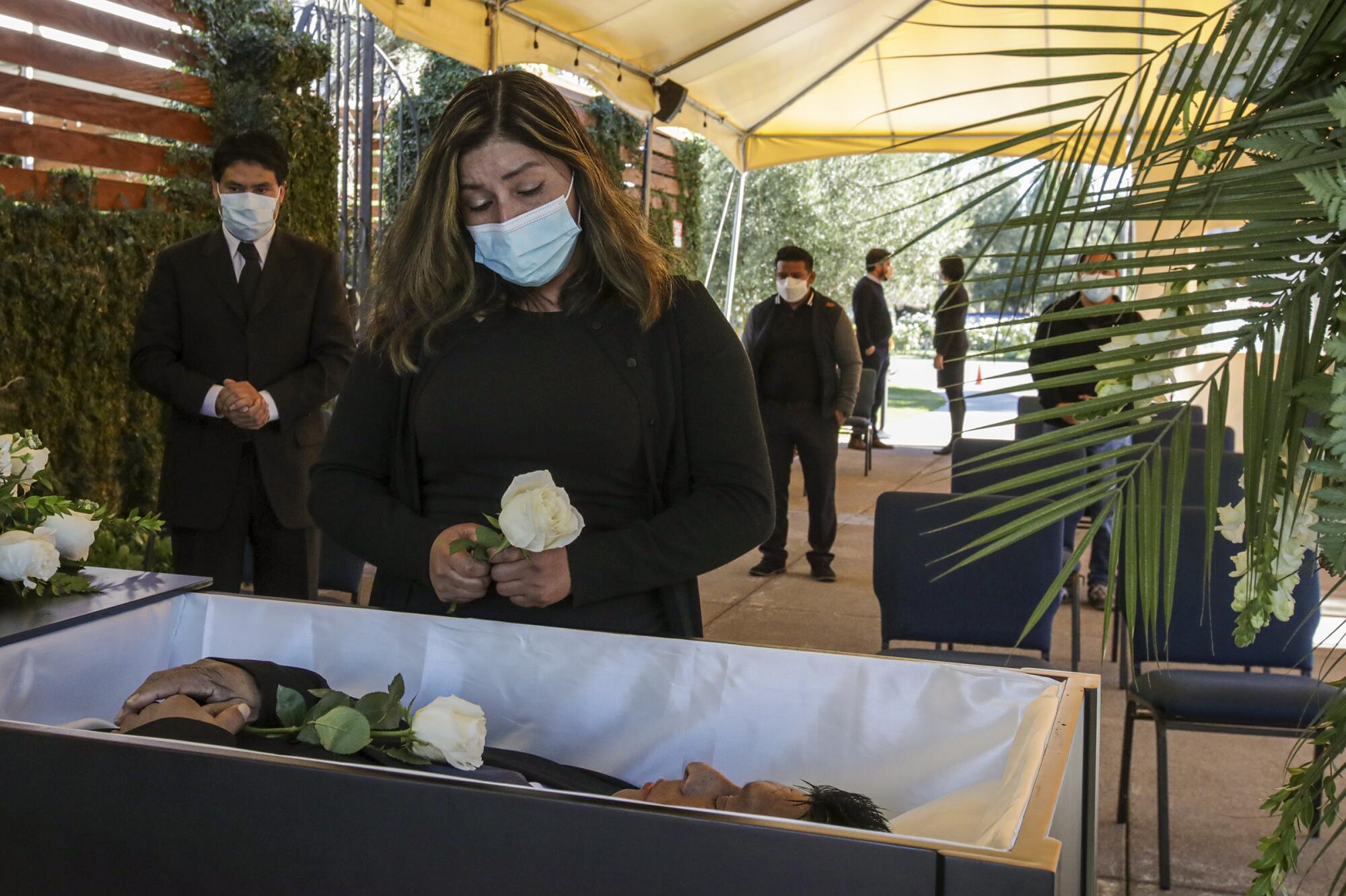A woman wearing a face mask holds a white rose beside a man's open casket as other mourners wait under an outdoor canopy