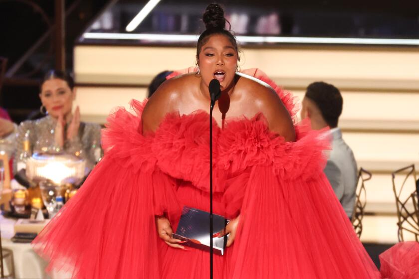 Lizzo, with a bun and a red poofy ball gown, accepts an Emmy behind a skinny microphone