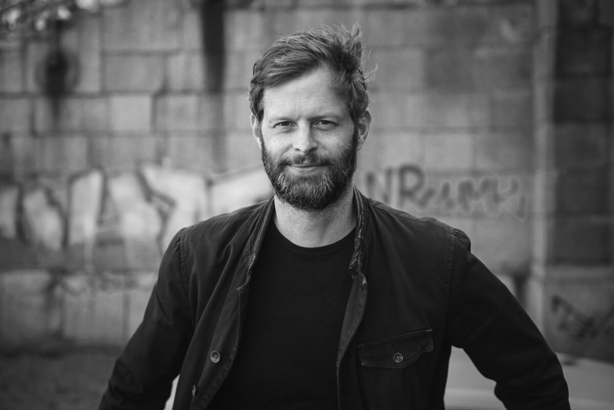 A man with a beard wearing black clothes in front of a brick wall with graffiti.