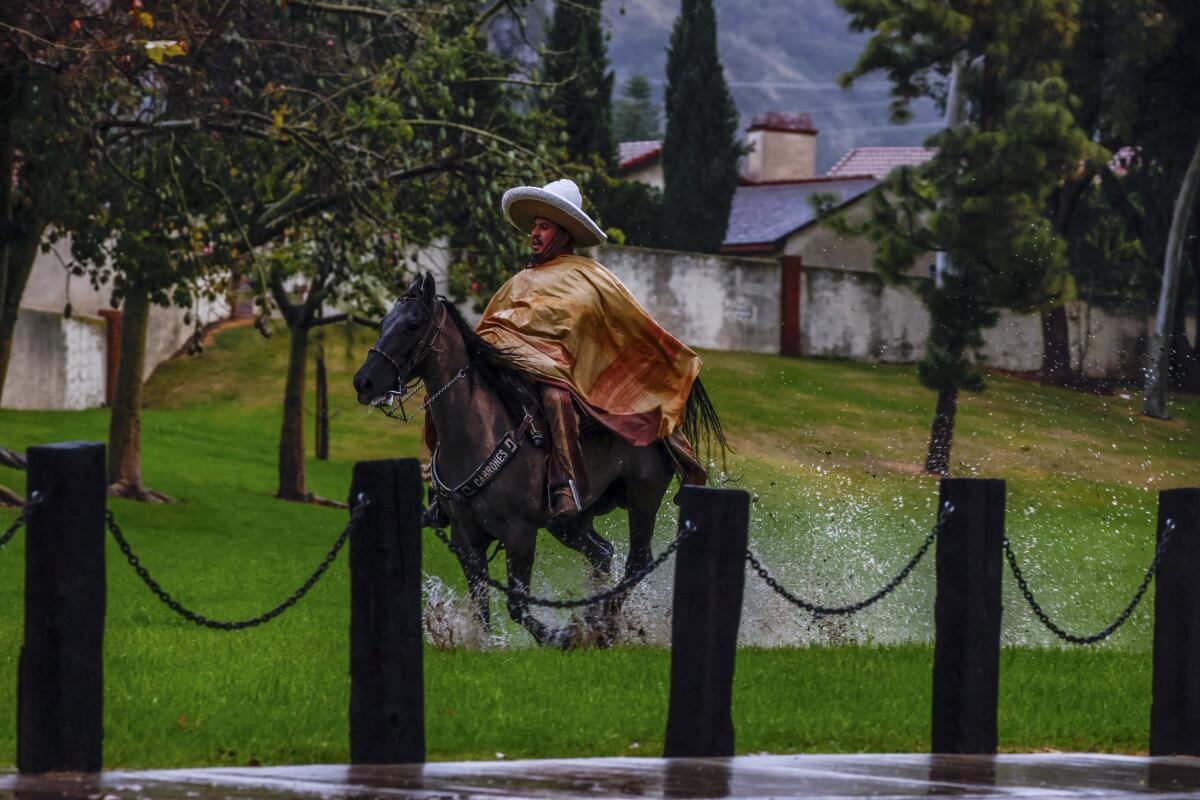 A man in a poncho and large hat rides a horse in the rain through soggy terrain, making a splash