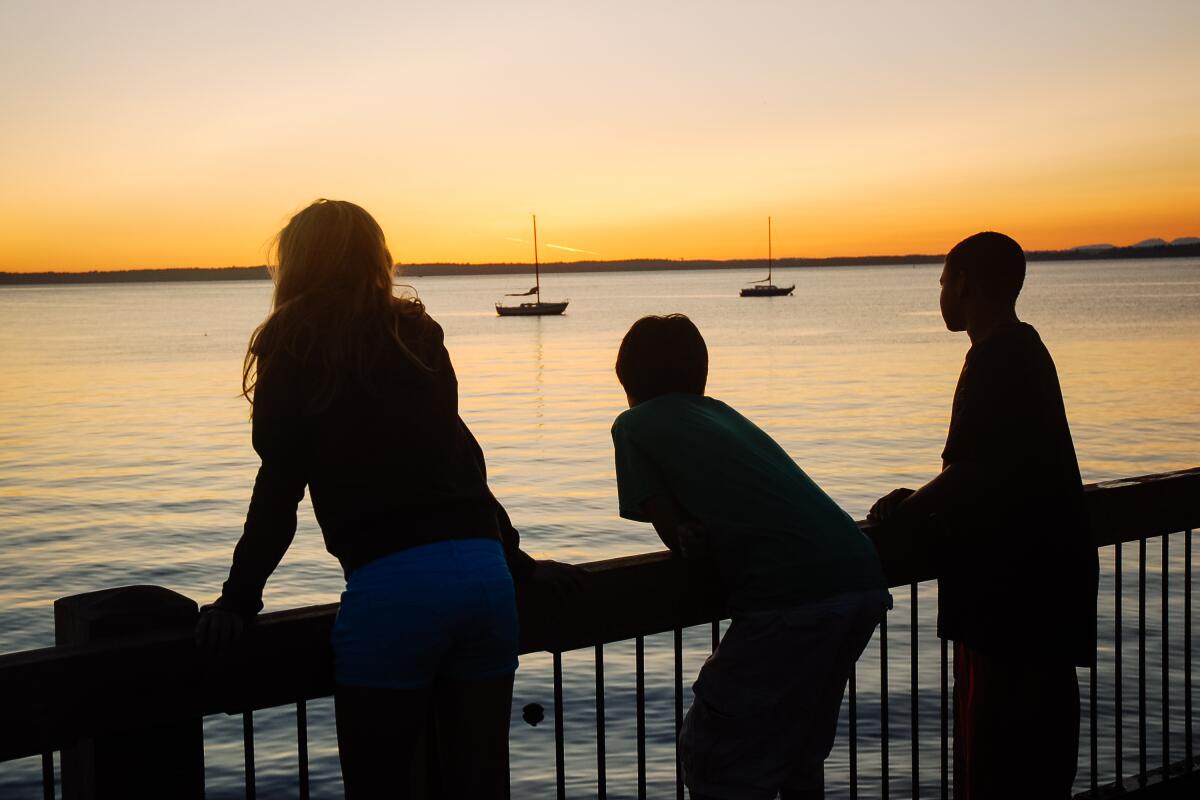 Drie mensen aftekenen tegen water met zeilboten bij zonsondergang