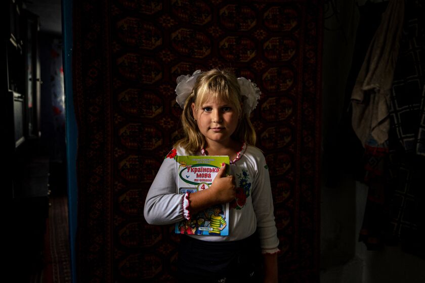 Standing in a dark room, lit by window light, a young blond girl with pigtails holds a schoolbook to her chest.