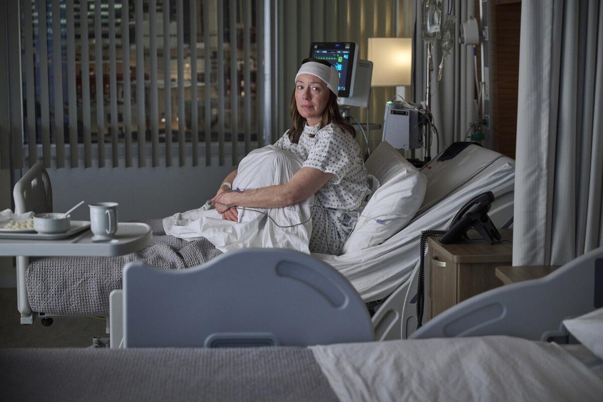 Uma mulher com uma bata de hospital e uma bandagem branca na testa está sentada na cama com os joelhos voltados para o peito.