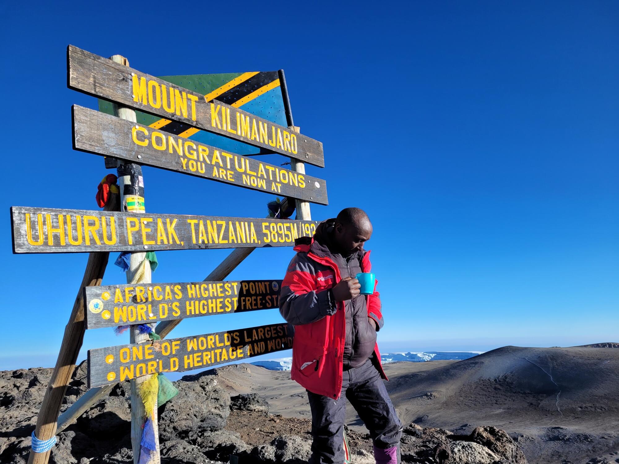 A man in a red jacket stands near a sign with five horizontal boards announcing the location, Uhuru Peak at Mt. Kilimanjaro