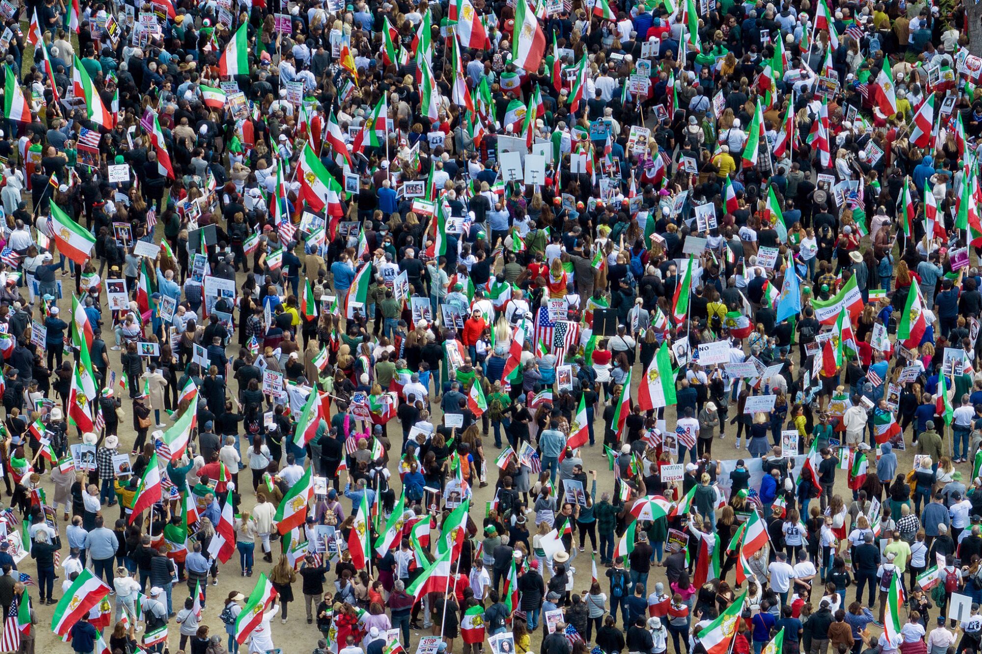 An aerial view of hundreds of small figures, many with Iranian and American flags, fill the entire frame. 