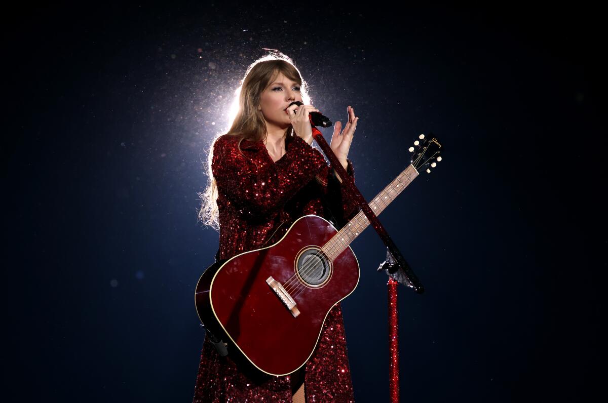 Taylor Swift performs onstage, singing into a microphone and with a guitar on a strap around her neck.
