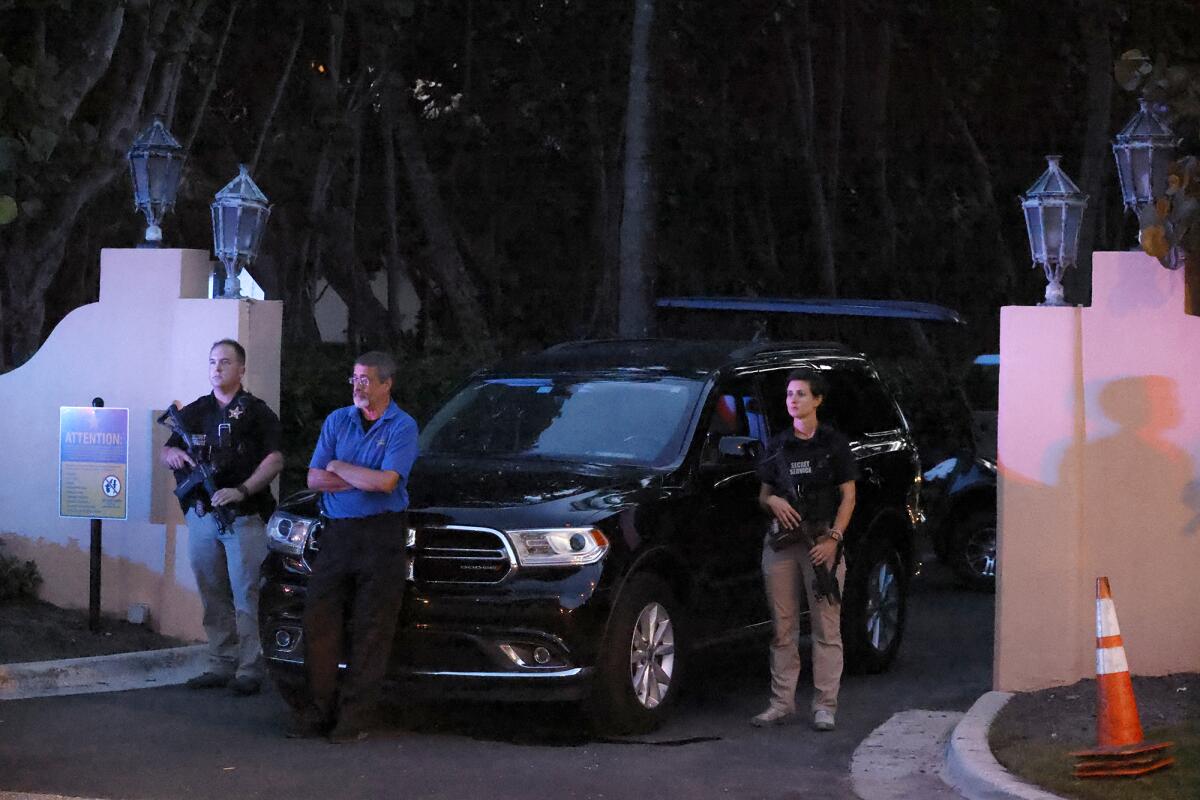 Three people, two clearly armed, standing by a black car at an entry gate to a property