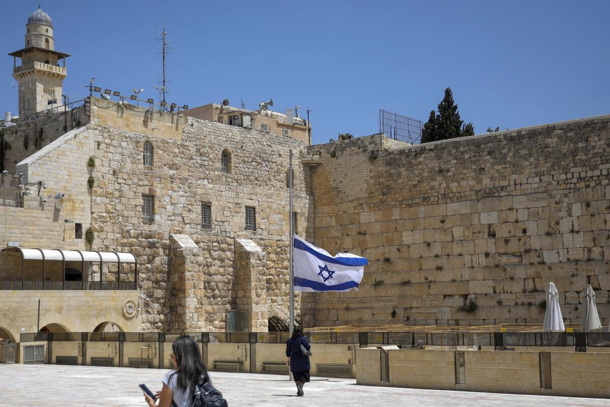 Israel's national flag is lowered to half-mast among buildings and pedestrians.