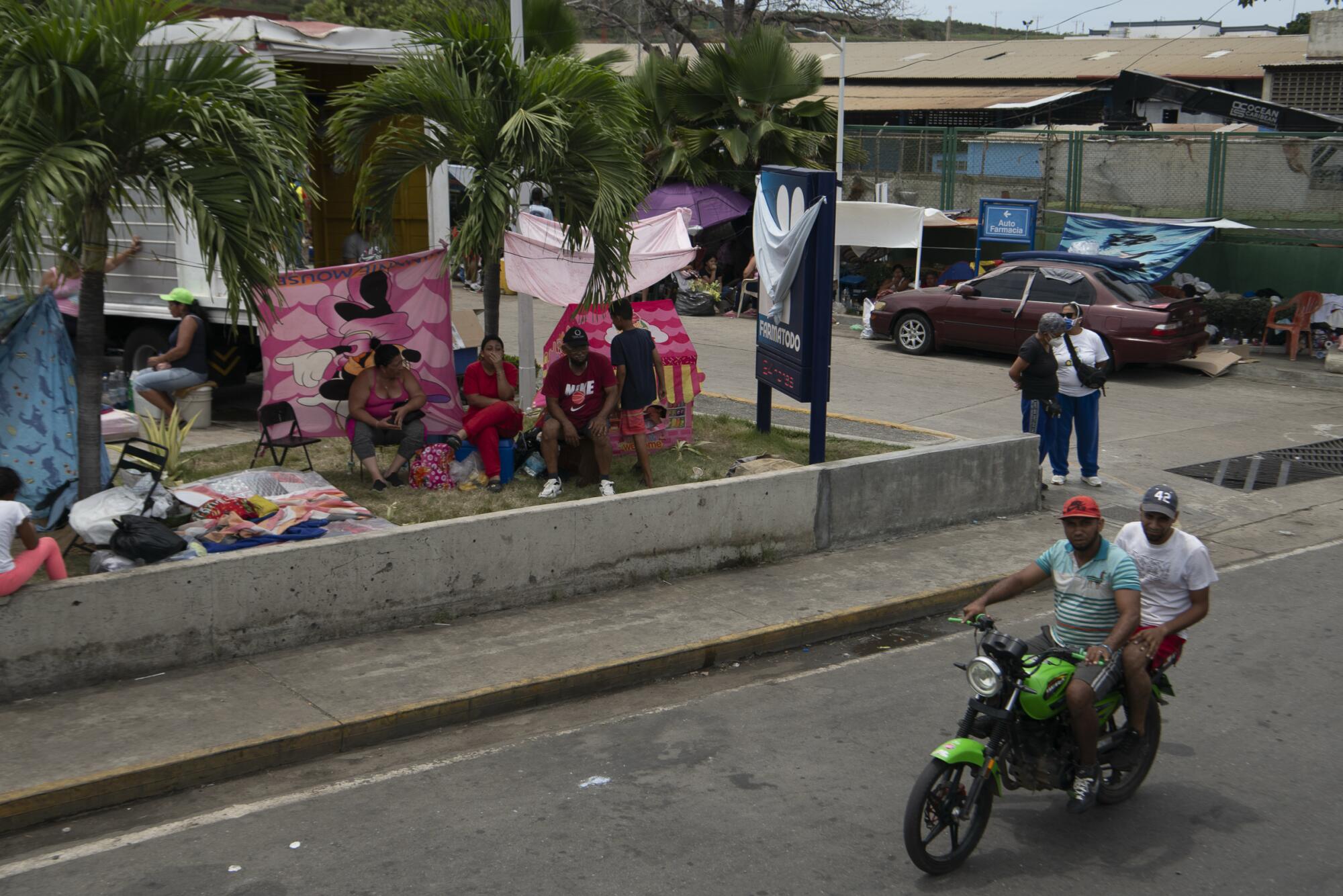 Two people ride a motorcycle past people sitting in a yard with their belongings