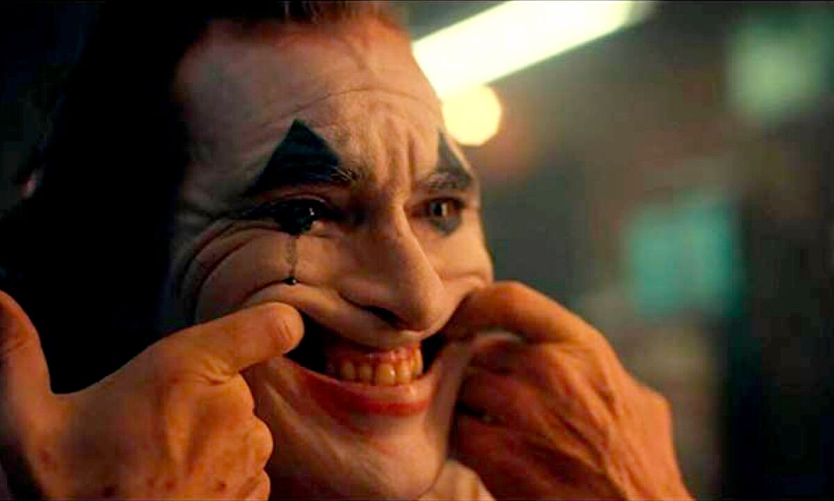 Joaquin Phoenix, with his index fingers hooked in his mouth, pulls his face into an exaggerated grin as the title character in "Joker."