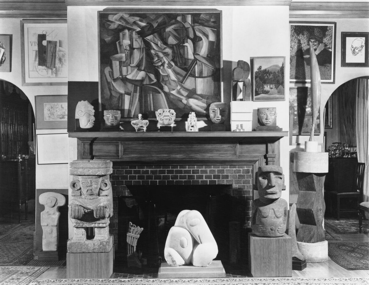 A black-and-white photograph shows paintings and sculpture surrounding a fireplace
