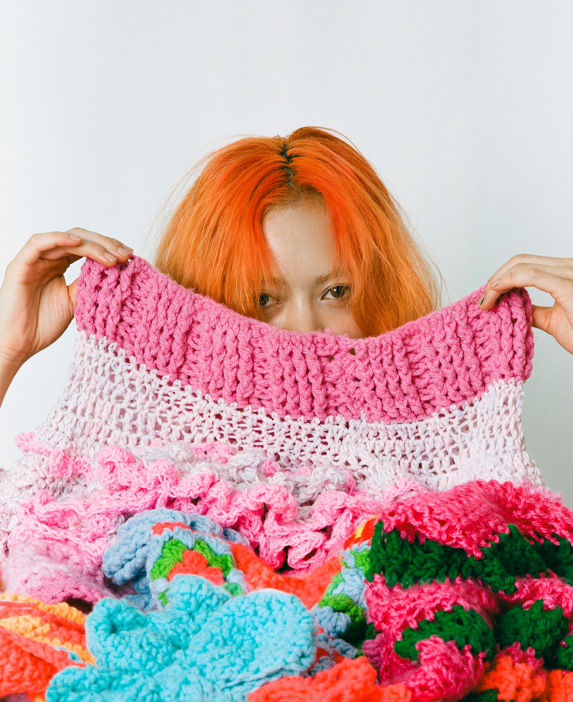 A woman holds up colorful crochet work, concealing half her face.