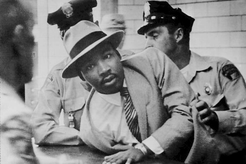 A Black man in a hat and suit and tie being jostled and handcuffed by white police.