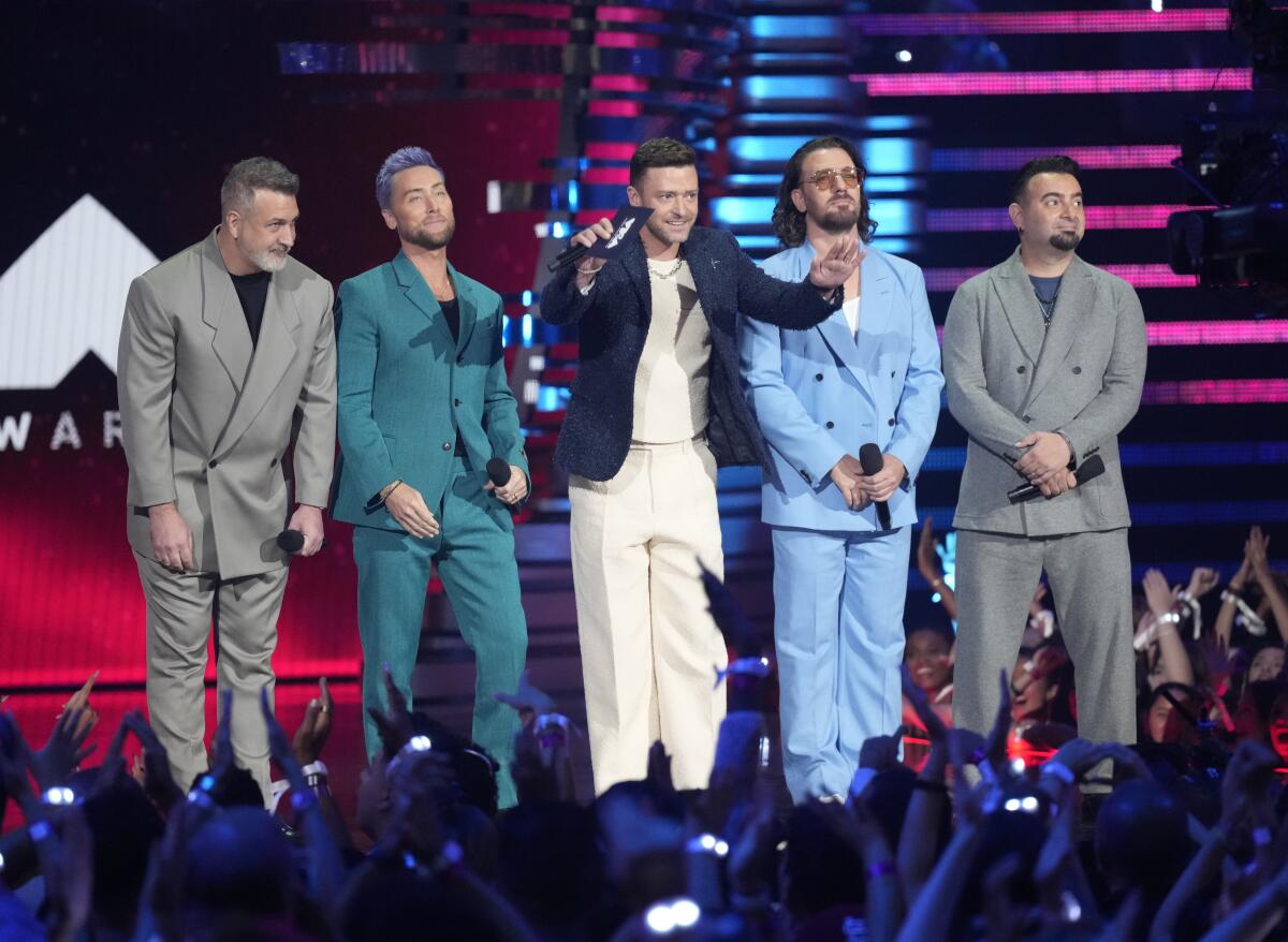 The five members of band NSYNC stand in a row onstage in suits as Justin Timberlake spreads his arms to quiet the audience