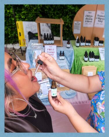 Jocelyn Herrera (L) opens her mouth to receive drops of CBD Mellow Out Tincture from Chelly XiCali of XiCali Products 