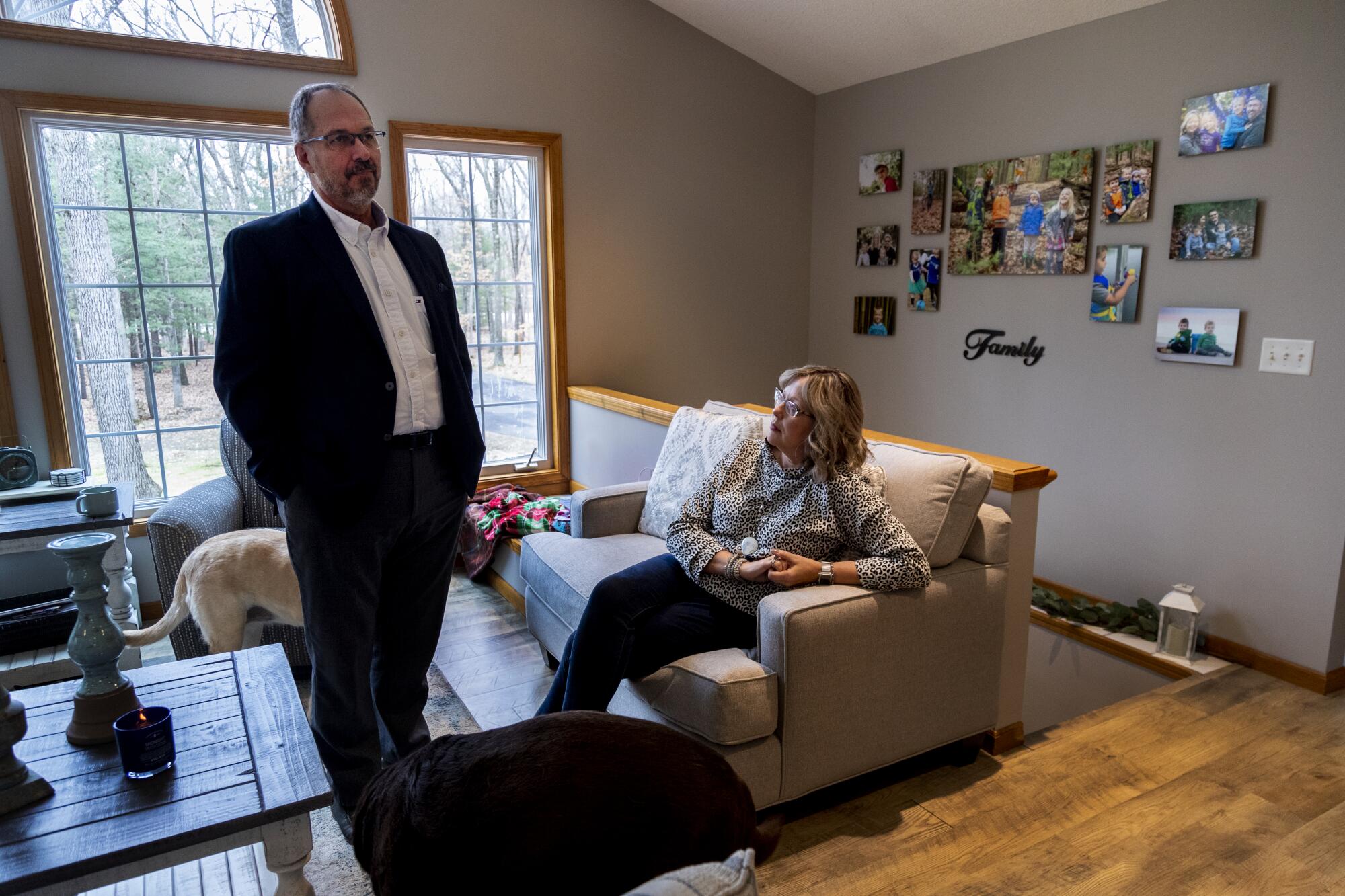 A man standing in a living room beside a seated woman, near a display of photos with the word "Family."