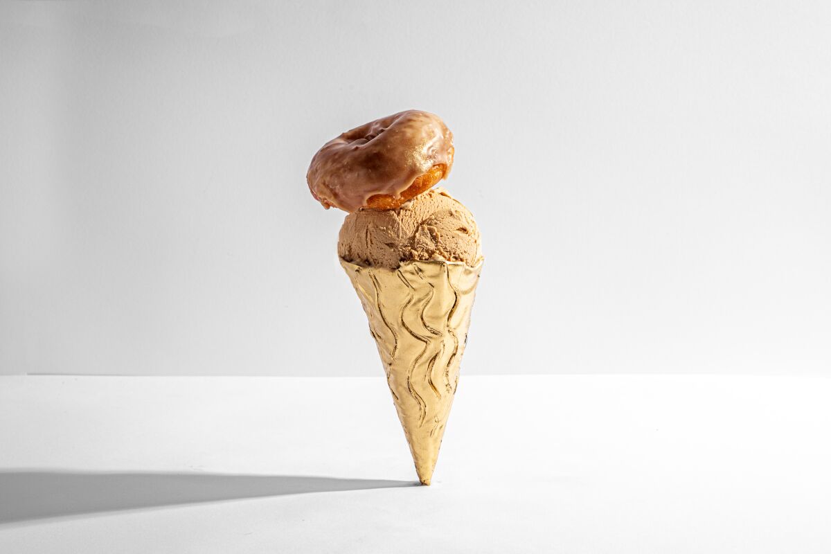 A doughnut-topped scoop of coffee gelato stands on the point of its gold-coated cone.