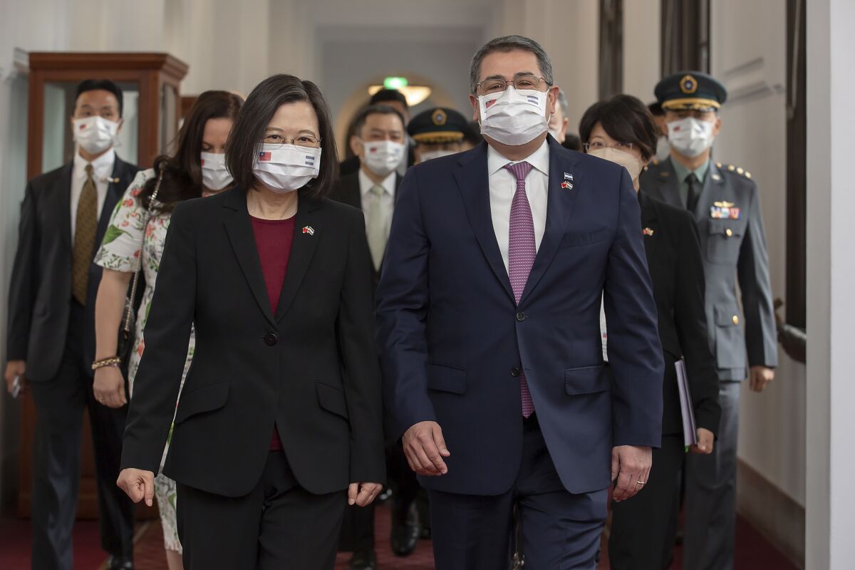 A woman and a man, both masked and in dark attire, walk in front of a group of people, also masked 