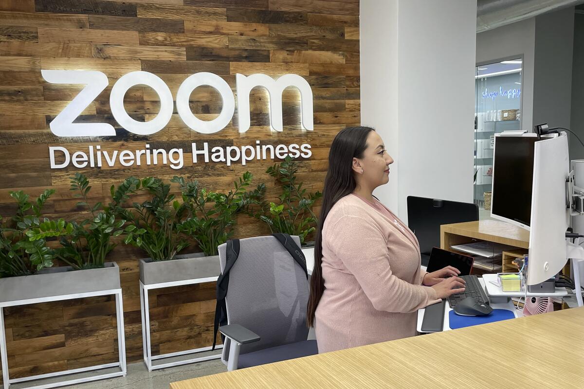 A woman is seated while working at a computer. On the wall behind her it says, "Zoom Delivering Happiness.".