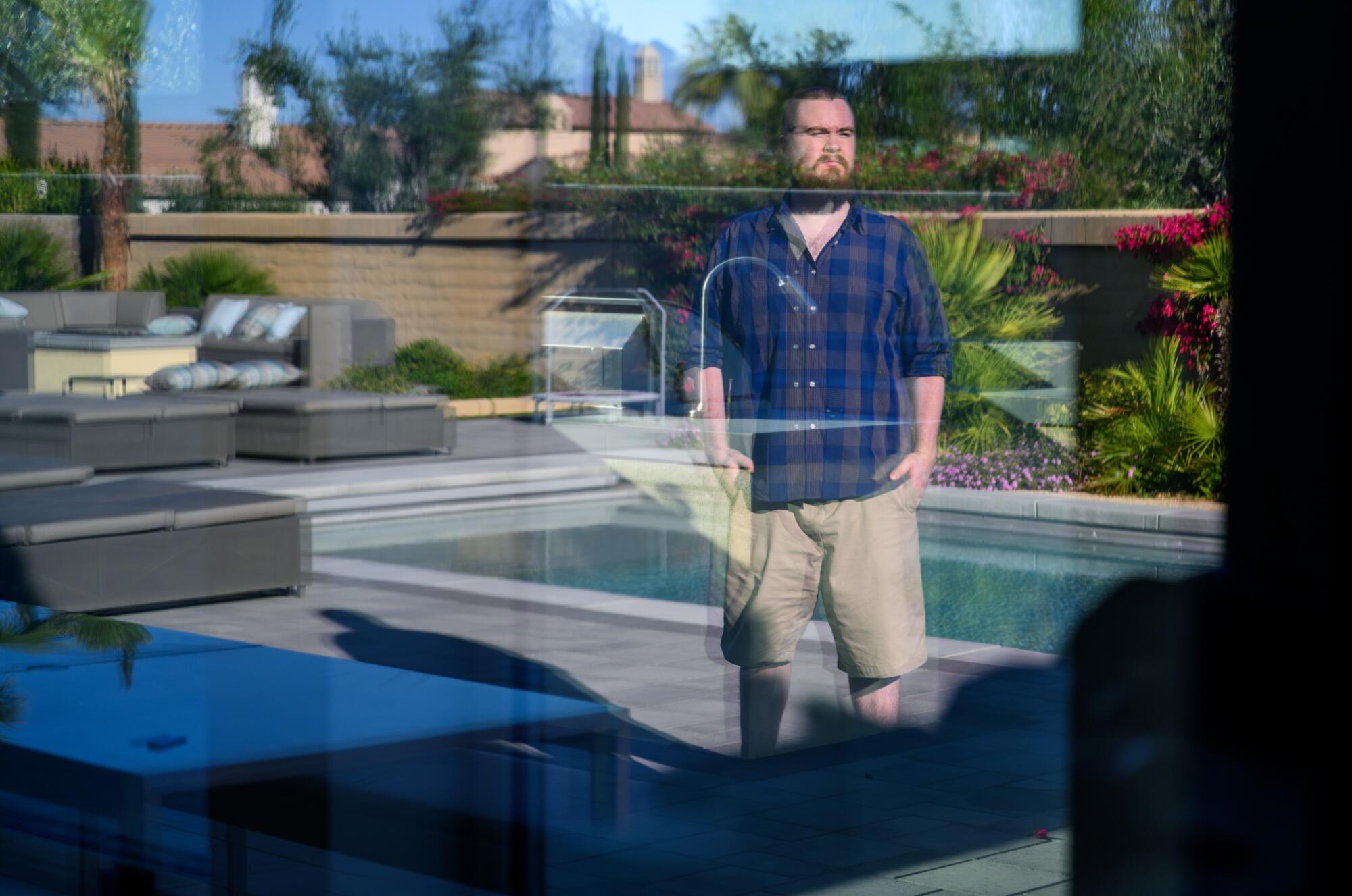 A man seen through a window stands outside near a swimming pool.