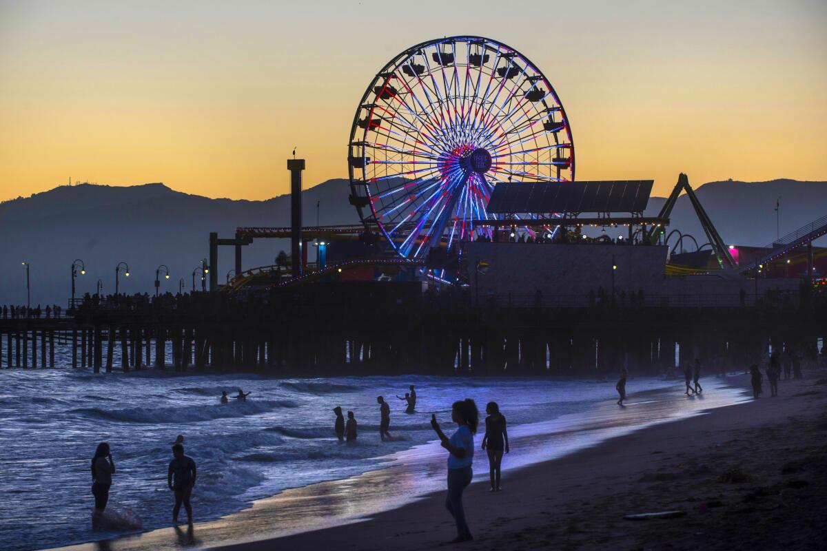 People stand in the water at the beach, with the Santa Monica Ferris wheel lighted up in the background.