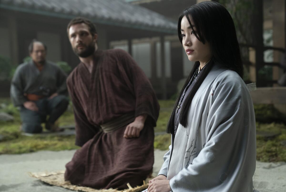 A man in a brown robe looks toward a woman in a white kimono.