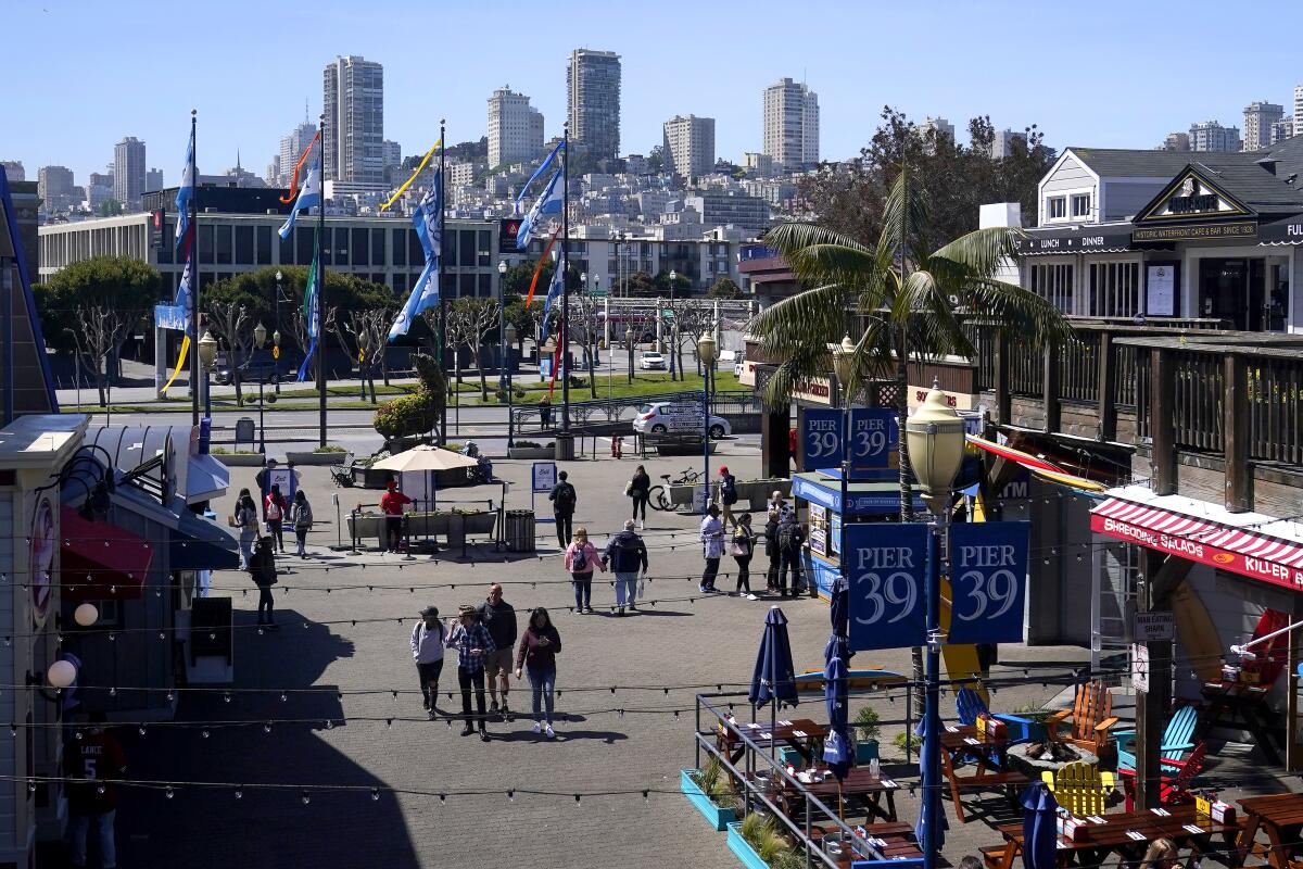 An elevated view of Pier 39. People walk about in between shops. High-rises and houses populate a small hill on horizon