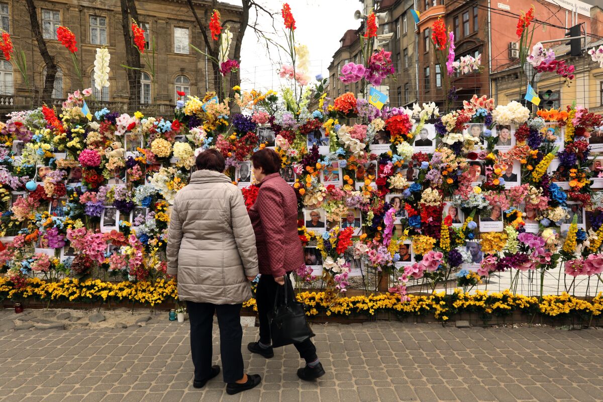 Two people stand looking at a wall of flowers and photos
