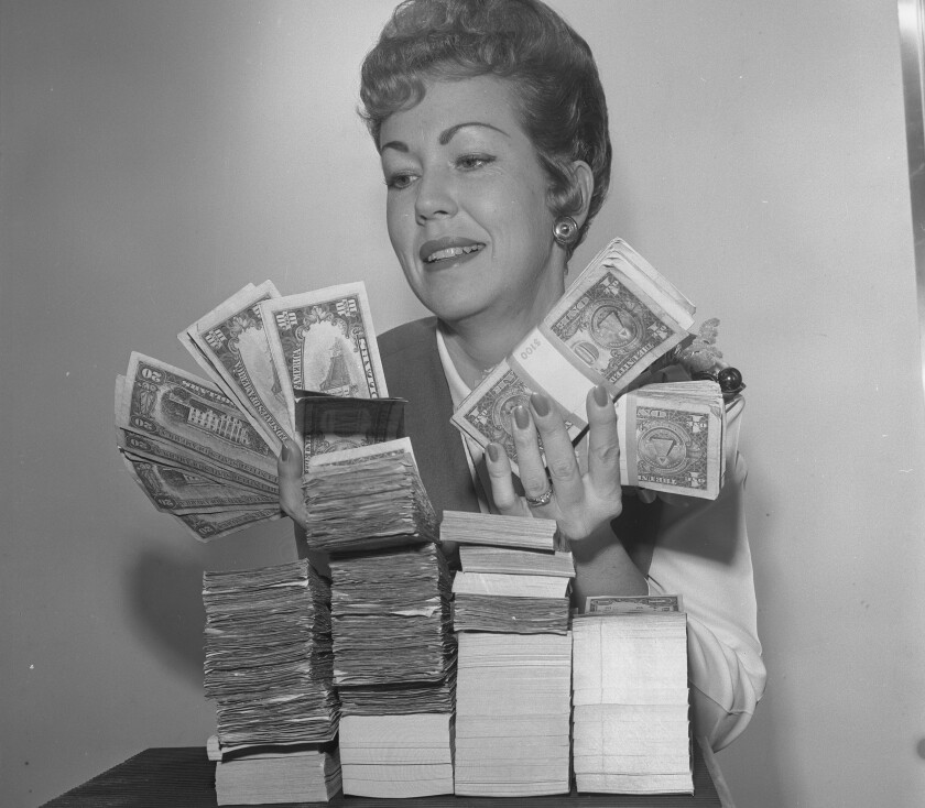A woman holds dollar, 10 and 20 bills in front of four small stacks of cash.