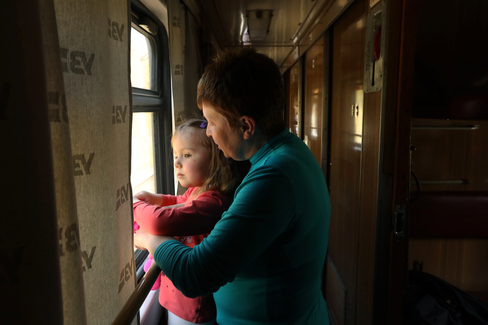 A woman in a green top and a girl in a pink shirt in front of her are looking out a window 