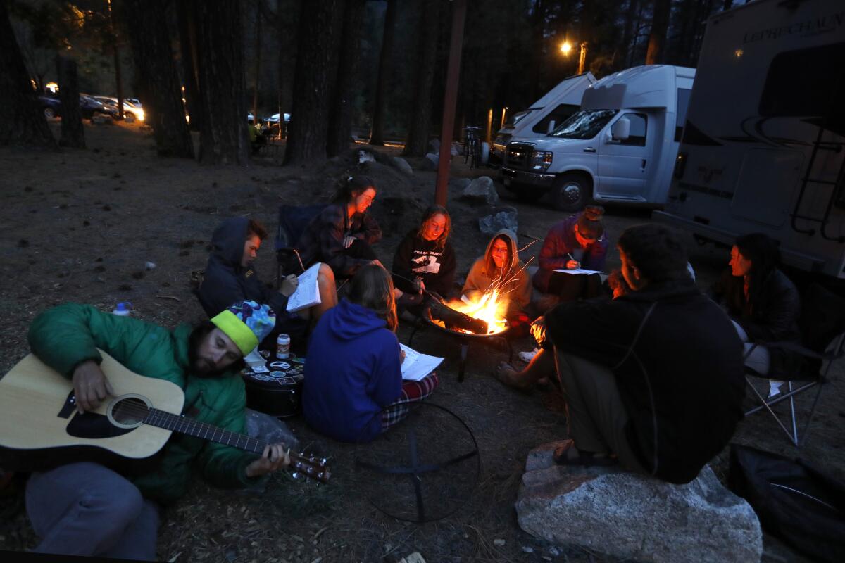 A group of students, including one with a guitar, sits around a campfire at a campground.