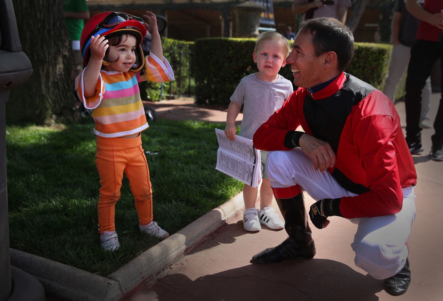 Valentina Pereira, 2, left, tries on her dad's jockey helmet, Tiago Pereira, right, before his race as Vincent Talamo, 2, watches while holding a racing program as Santa Anita resumes racing at Santa Anita Horse Park in Arcadia, Calif., on March 29, 2019. (Allen J. Schaben / Los Angeles Times)