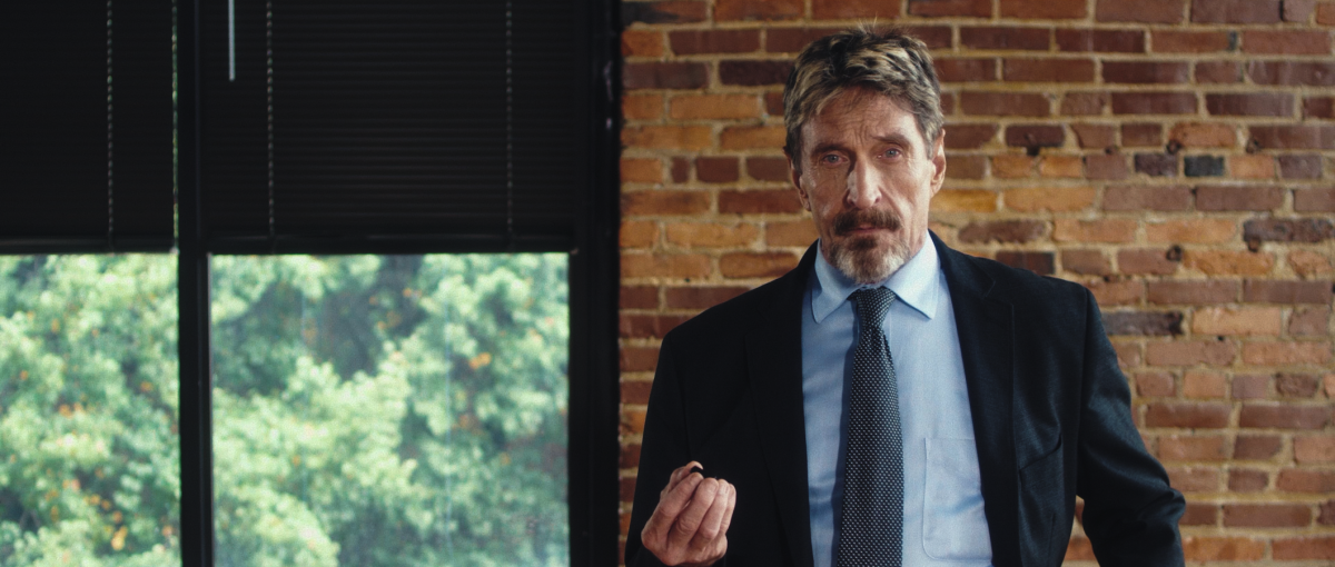 A man with a beard and mustache, wearing a suit, stands before a window that's in a brick wall.