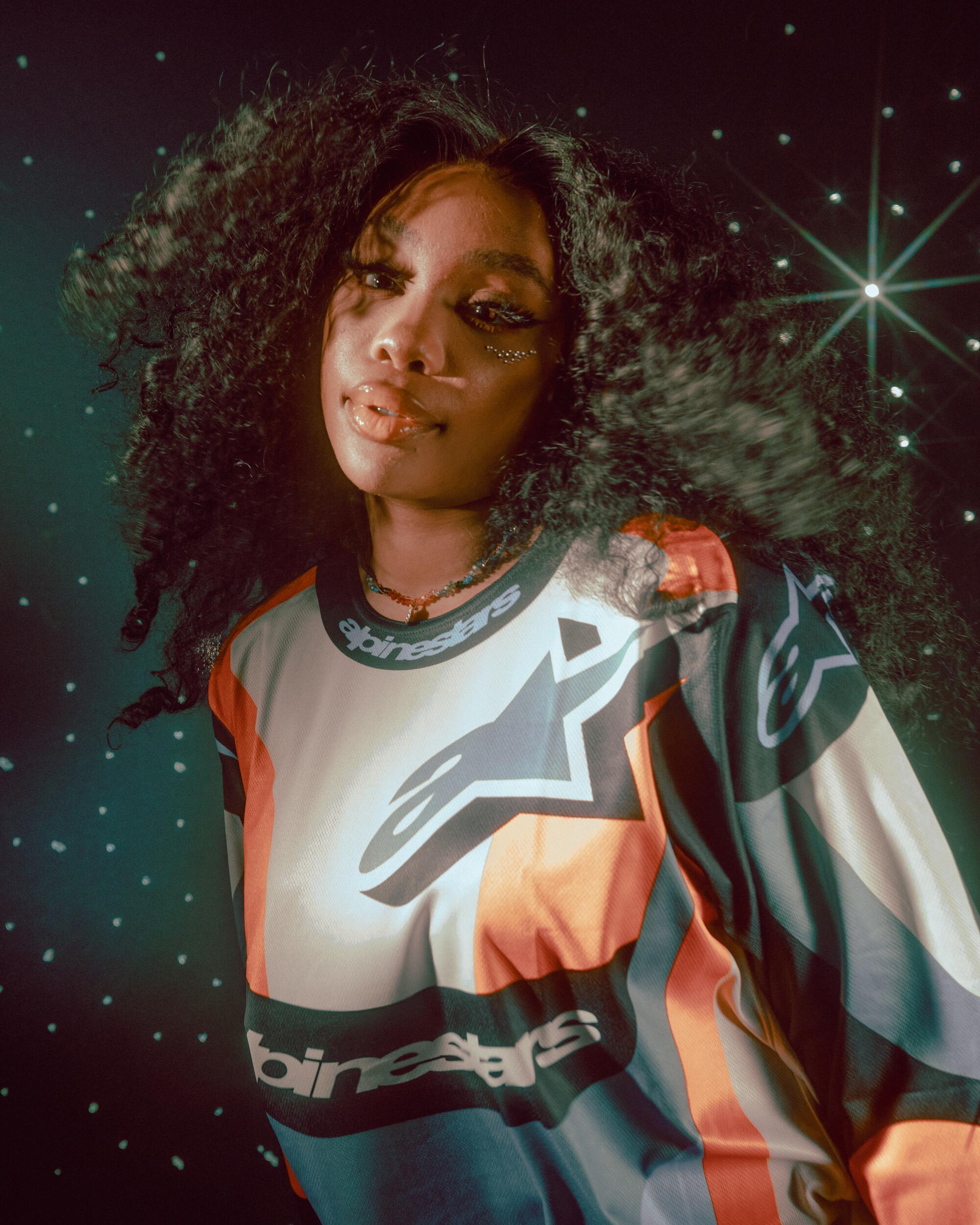 SZA wears a colorful shirt and stands in front of a celestial backdrop. 