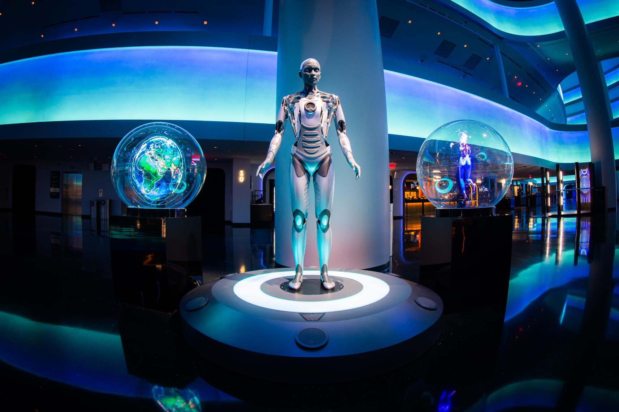 A robot with a woman's face and a mechanical body standing between two glass spheres in an atrium illuminated in blue