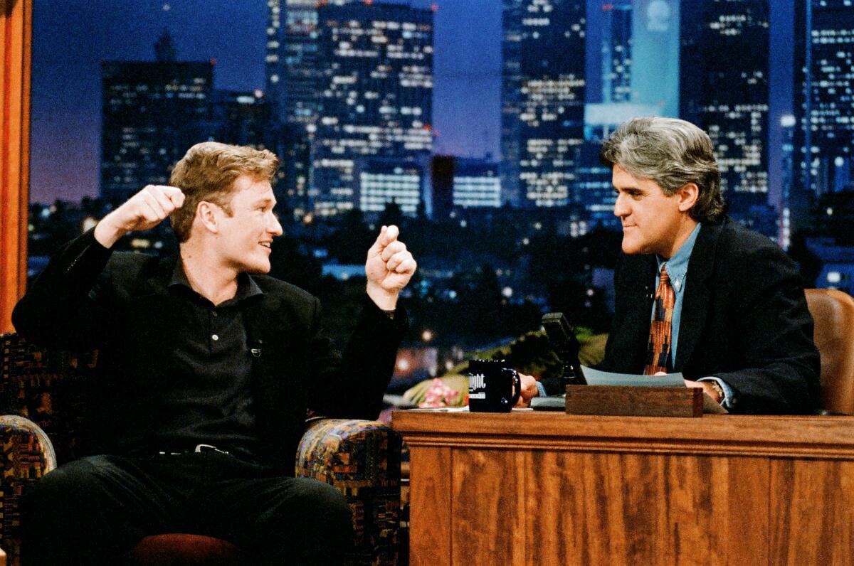 Conan O'Brien holds his arms up as he sits in chair next to Jay Leno, who is behind his desk.
